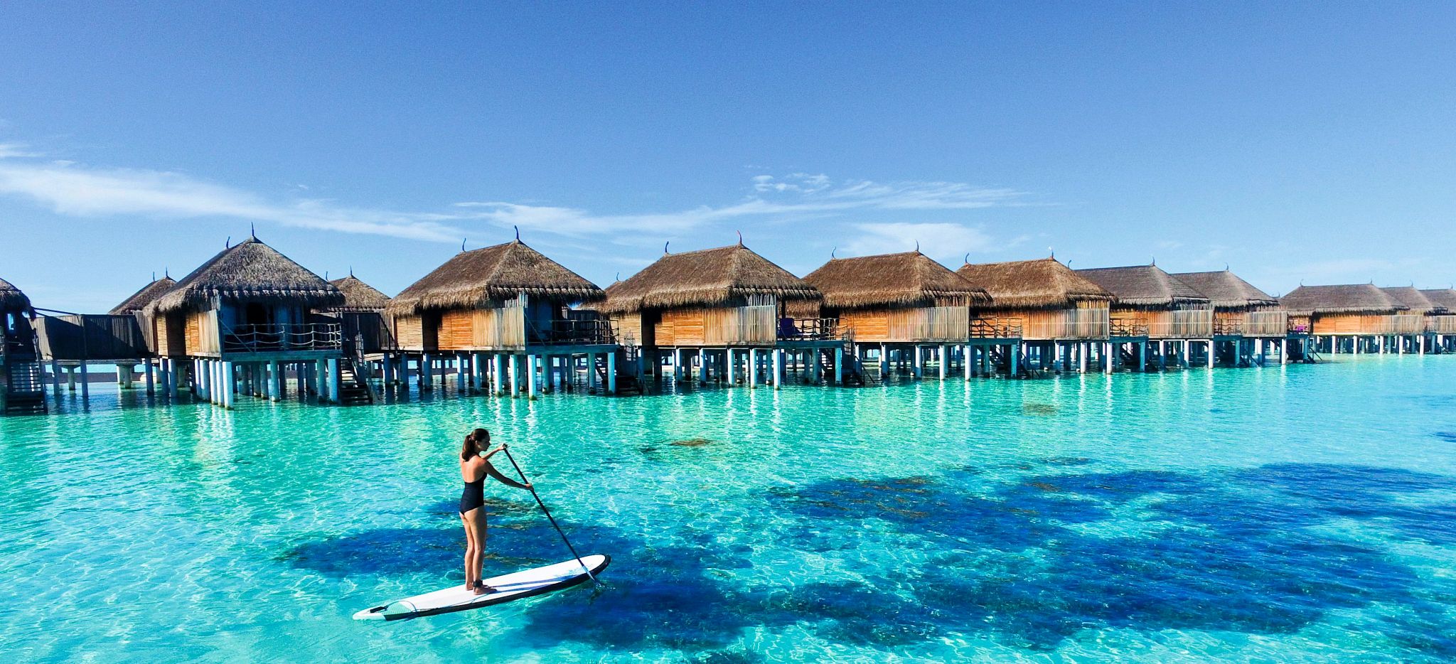 Eine Frau paddelt auf eine SUP an den Wasservillen des Constance Moofushi entlang